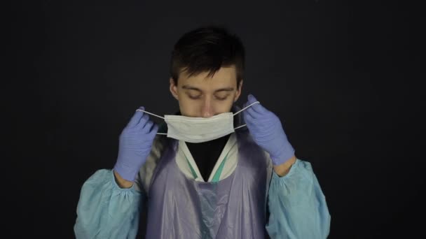 Ένας νεαρός καυκάσιος γιατρός που έχει δυσκολία στην αναπνοή, βήχα. Φορώντας ιατρικά προστατευτικά ρούχα, κοστούμι, μάσκα, γάντια. Απομονωμένο μαύρο φόντο. Coronavirus, Covid19. — Αρχείο Βίντεο