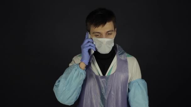 A young caucasian doctor calling someone on the cell phone. Wearing protective medical suit. Isolated dark background. Coronavirus. — Stock Video