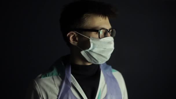En kaukasisk man som har svårt att hälla på sig en medicinsk mask. Skrattar och ler. Coronavirus. Mörk bakgrund. — Stockvideo