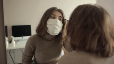 Koruyucu maskeli genç bir kız karantinada otururken aynanın önünde makyaj yapıyor. Coronavirüs. COVID-19