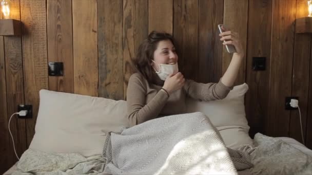 Een mooi beschermend gemaskerd meisje praat thuis aan de telefoon tijdens de quarantaine. Coronavirus. COVID-19 — Stockvideo