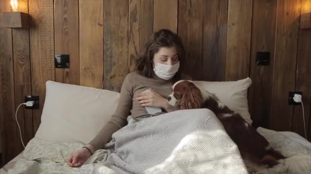 Ein junges Mädchen in Schutzmaske spielt mit ihrem lustigen Hund, der in der Isolation im Bett liegt. Coronavirus. COVID-19 — Stockvideo