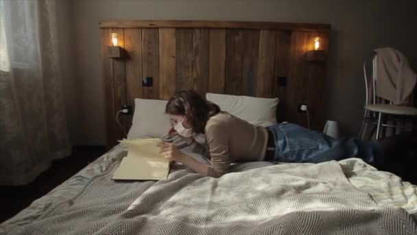 A young European girl in her room lies in a protective mask and turns over a book during quarantine.Coronavirus. COVID-19 — Stock Video