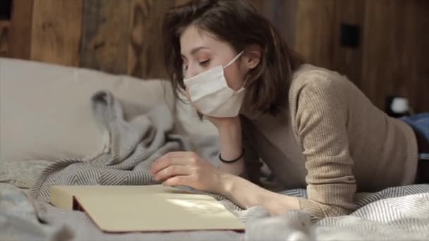 Κοντινό πλάνο ενός νεαρού κοριτσιού με προστατευτική μάσκα που ψεύδεται και διαβάζει ένα βιβλίο.Coronavirus. COVID-19 — Αρχείο Βίντεο
