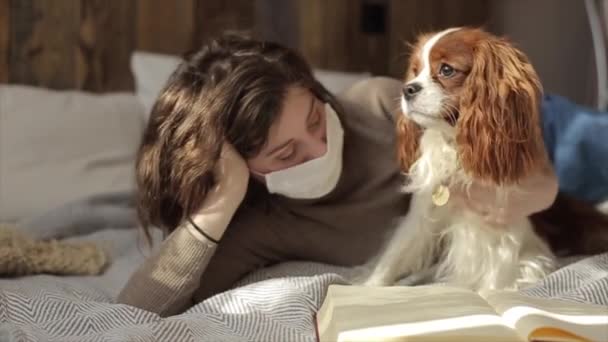 Lento movimiento de una joven chica enmascarada protectora y su lindo cachorro en la cama al lado de un libro durante la cuarentena. COVID-19 — Vídeos de Stock