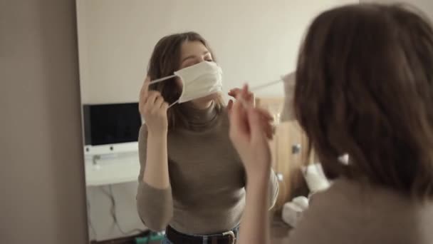 Молодая красивая девушка надевает защитную маску перед зеркалом и выпрямляет волосы. Coronavirus.COVID-19 — стоковое видео