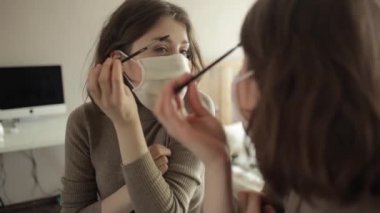 Tıbbi maskeli genç bir kadın aynanın önünde göz makyajı yapıyor. Coronavirus. COVID-19.