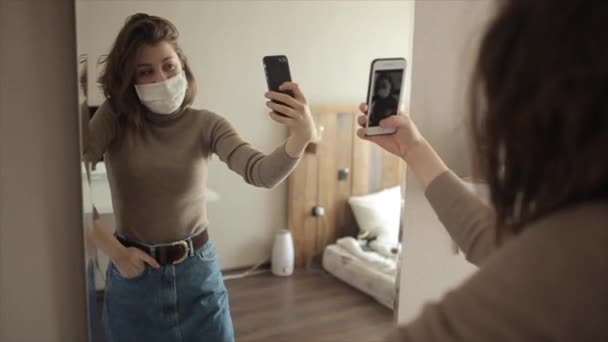 Carino ragazza si trova di fronte a uno specchio protettivo mascherato e si fa un selfie. Coronavirus. COVID-19 — Video Stock