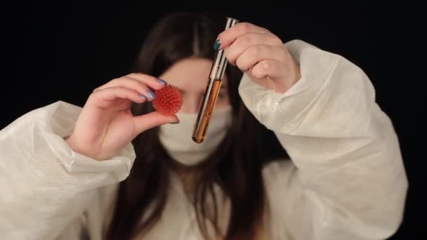 Девушка в защитном костюме и медицинской маске показывает камере лабораторные колбы с жидкой и коронавирусной мини-моделью вируса COVID-19 — стоковое видео