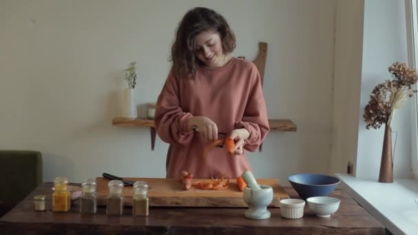 Милая молодая девушка чистит морковь на доске для резки овощей на столе со специями на кухне — стоковое видео