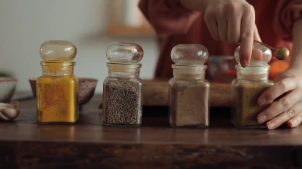 En ung flicka tar en sked kryddor från burkar som står på ett bord i köket. Närbild — Stockvideo