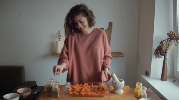 Young beautiful girl in the kitchen puts spices in a mortar to grind them and sprinkle the dish with carrots for baking — Stock Video