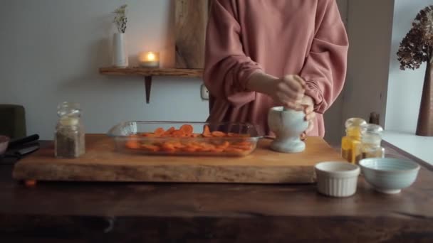 Dziewczyna przy kuchennym stole młóci przyprawy w zaprawie obok szklanej miski posiekanych marchewek. Zbliżenie — Wideo stockowe