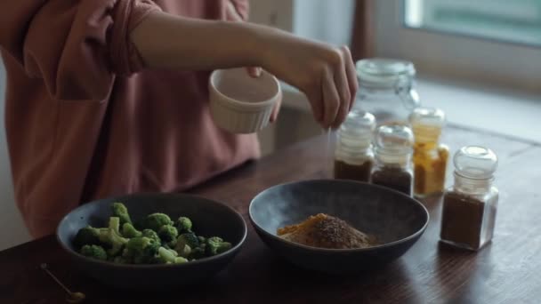Fiatal lány keveredik egy tál fűszerek sóval brokkoli a konyhaasztalon konzervek fűszerek. Közelkép — Stock videók