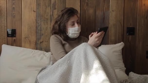 Νεαρό όμορφο κορίτσι κάθεται στο κρεβάτι σε μια προστατευτική μάσκα με ένα τηλέφωνο κατά τη διάρκεια της αυτο-απομόνωσης. Ιός του κερατοειδούς. COVID-19 — Αρχείο Βίντεο