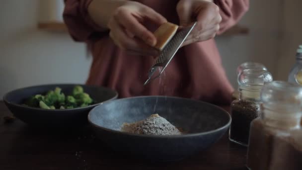 La muchacha frota el queso duro en un pequeño rallador en un tazón con especias para cocinar brócoli. Primer plano — Vídeo de stock