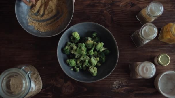 Chica esparcir el brócoli con una mezcla de especias de un tazón con las manos en la mesa de la cocina. Vista desde arriba — Vídeo de stock