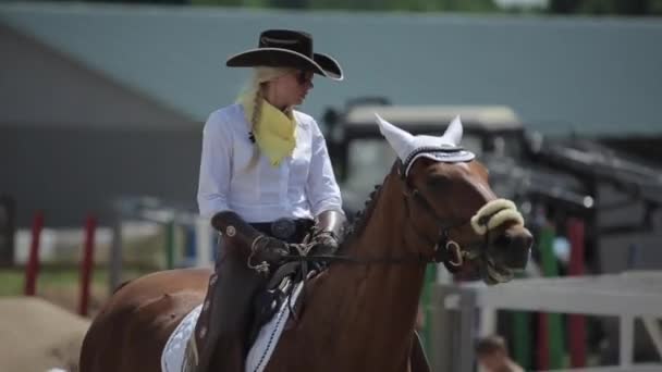 Minszk, Fehéroroszország - 2019. július 19.: Egy gyönyörű fiatal szőke cowboy jelmezben és sötét szemüvegben lassan lovagol a lován. Közelkép — Stock videók