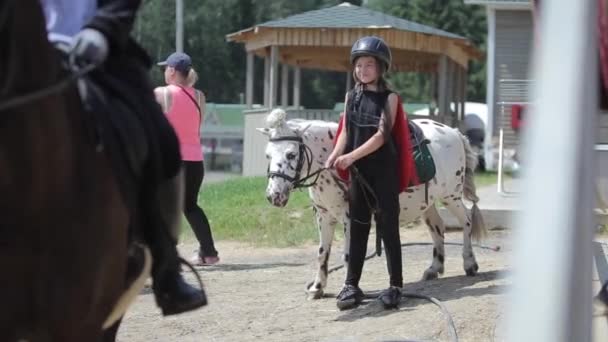 Mińsk, Białoruś - 19 lipca 2019: Mała piękna dziewczynka w czarnym garniturze i kasku stoi obok kucyka i uśmiecha się — Wideo stockowe