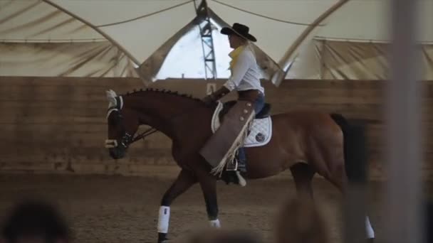 Minsk, Bielorrússia - 19 de julho de 2019: Jovem loira bonita em um traje de cowboy na sela de um cavalo calmamente andando em um rancho coberto de arena. Movimento lento — Vídeo de Stock