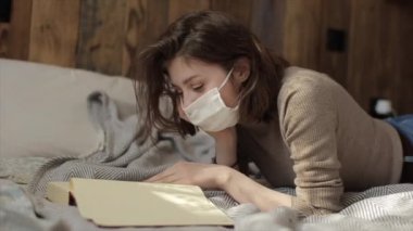 Korumacı maskeli genç bir kızın yalan söyleyip kitap okuduğu fotoğraf. Coronavirus. COVID-19