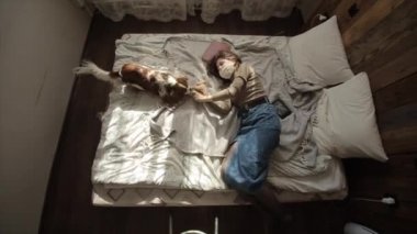 Koruyucu maskeli genç bir kadın kendini tecrit ederken köpeğiyle yatakta oynar. Yukarıdan bak. Coronavirüs. COVID-19