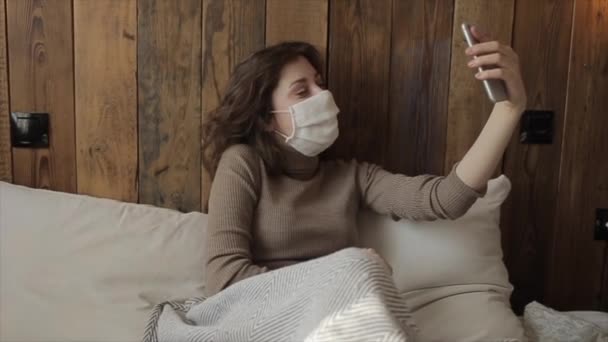 Молодая красивая девушка сидит в постели в защитной маске с телефоном и делает селфи во время самоизоляции. Коронавирус. COVID-19.Карантинная активность — стоковое видео