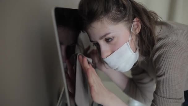 Uma jovem em casa em quarentena em uma máscara médica limpa cuidadosamente o computador com um desinfetante. Coronavirus.COVID-19 — Vídeo de Stock