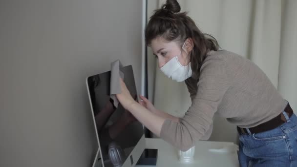 En ung flicka hemma i karantän i en medicinsk mask torkar försiktigt datorn med ett desinfektionsmedel. Coronavirus.COVID-19 — Stockvideo