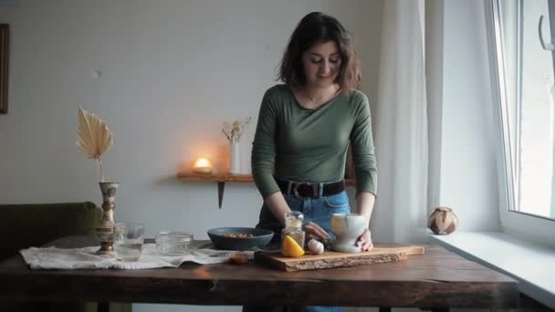 A cute young girl in her kitchen puts spices from a mortar into a bowl with chickpeas for cooking traditional hummus — Stock Video