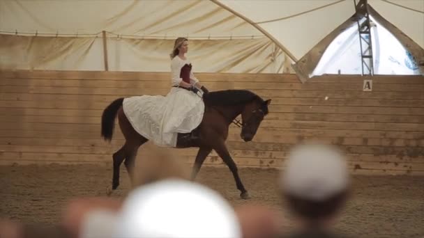 Minsk, Bělorusko - 19. července 2019: Kůň pomalu kráčí po ranči s krásnou mladou dívkou v sedle v dlouhých bílých šatech — Stock video