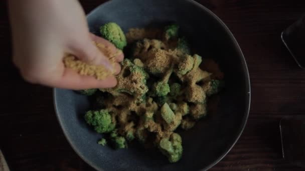 A lány brokkolit szórt fűszerek keverékével egy tálból, kezével a konyhaasztalnál. Kilátás felülről — Stock videók