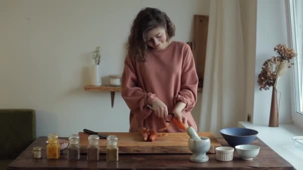 Милая молодая девушка чистит морковь на доске для резки овощей на столе со специями на кухне — стоковое видео