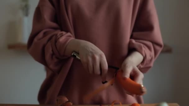 Крупним планом руки дівчини, яка очищає моркву спеціальним ножем на стільниці на кухні в її квартирі — стокове відео