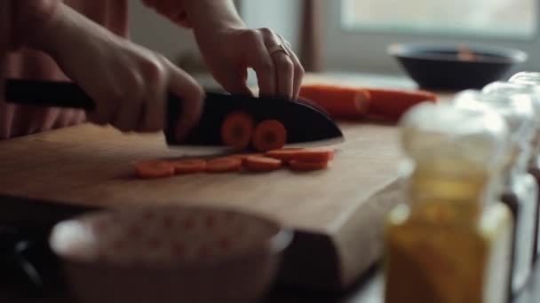 Das Mädchen schneidet mit einem großen Messer auf der Arbeitsplatte in ihrer Küche Möhren im Kreis. Nahaufnahme — Stockvideo