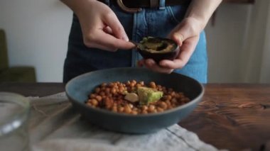 Genç bir kız evde geleneksel humus yapmak için içinde nohut olan bir kâseye avokado ekler. Yakın plan.