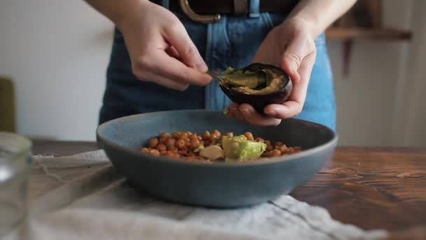 A young girl adds avocado in a bowl with chickpeas to make traditional hummus at home. Close-up — Stock Video