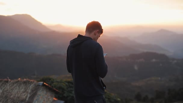 Een man kijkt naar zijn telefoon terwijl hij een prachtig uitzicht heeft op het berglandschap bij zonsondergang, zonsopgang. — Stockvideo