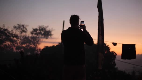 Uma silhueta de um homem a fotografar o pôr-do-sol, o nascer do sol. Vila, secagem de roupa em uma linha ao ar livre . — Vídeo de Stock