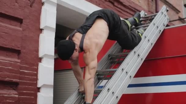 Ένας νεαρός μυώδης άντρας περπατάει με τα χέρια του κάτω από μια σκάλα που συνδέεται με ένα πυροσβεστικό όχημα. Κοντινό πλάνο — Αρχείο Βίντεο