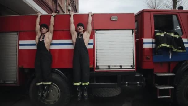 Zwei muskulöse Feuerwehrmänner schwingen sich synchron auf das Dach eines Feuerwehrfahrzeugs. Nahaufnahme — Stockvideo
