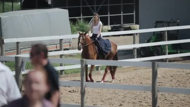 Minsk, Bielorrusia - 19 de julio de 2019: Hermosa joven rubia montando a caballo en un vestido largo monta lentamente en un corral en un rancho de caballos — Vídeo de stock