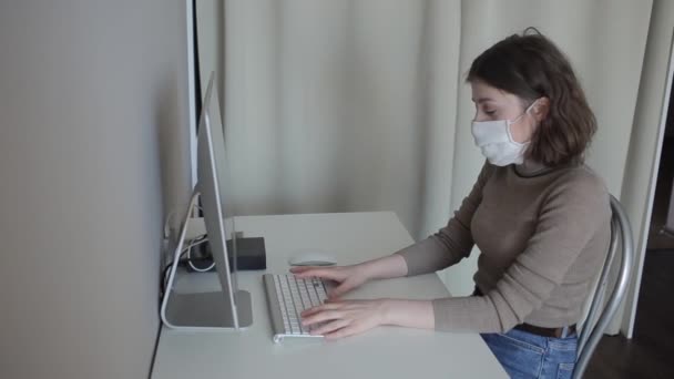 Chica joven con una máscara protectora hace un calentamiento sentado en una mesa con una computadora en cuarentena en casa.Coronavirus.COVID-19 — Vídeo de stock