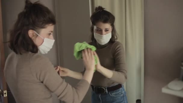 Une jeune fille européenne portant un masque de protection essuie un miroir avec un antiseptique assis à la maison en quarantaine. COVID-19 — Video