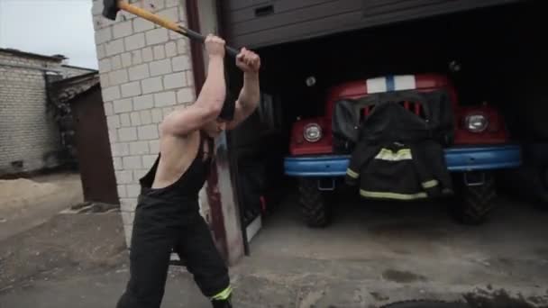 Männer bei Übungen und Workout. Zwei muskulöse junge Feuerwehrleute schlugen mit einem Vorschlaghammer auf den Reifen eines Feuerwehrfahrzeugs in einer Garage ein. Zeitlupe. Nahaufnahme — Stockvideo