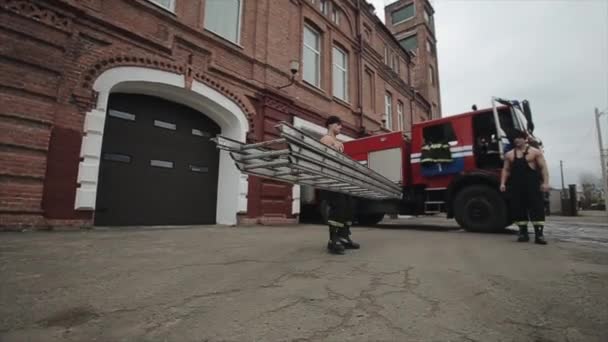 Dos jóvenes bomberos musculosos están entrenando con una gran escalera de metal al aire libre en el fondo de un camión de bomberos — Vídeos de Stock