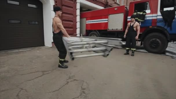 Hombres haciendo ejercicios y entrenamiento.Un joven musculoso tuerce una larga escalera de metal y otro hombre salta a través de ella contra el fondo de un camión de bomberos. Primer plano — Vídeo de stock