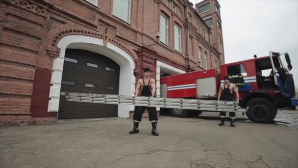 Men doing exercises and workout. Young muscular fireman jumps over a long metal ladder that another fireman twists in his hands against the background of a fire truck — Stock Video