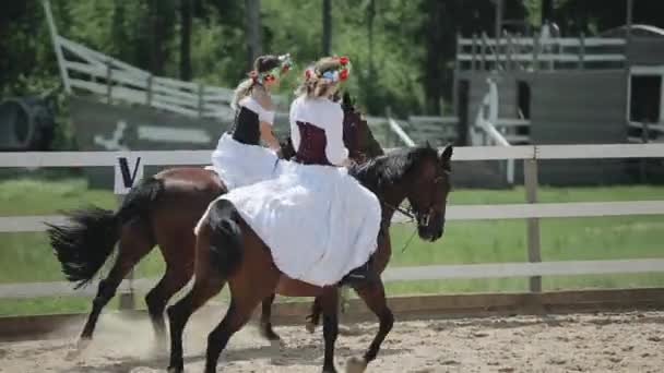 Minsk, Vitryssland - 19 juli 2019: Två unga flickor i vackra klänningar och med kransar på huvudet rider bredvid varandra till häst på en ranch — Stockvideo