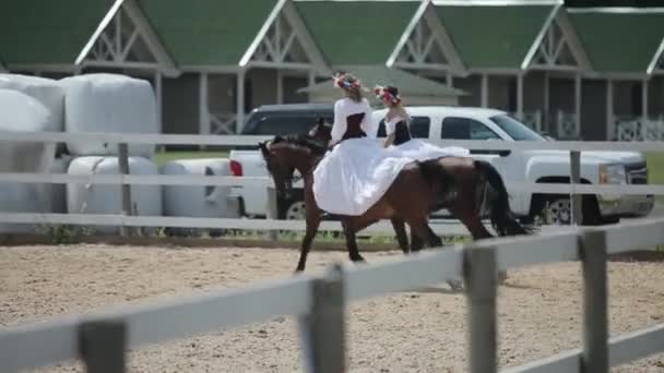 Minsk, Vitryssland - 19 juli 2019: Två unga flickor i vit medeltida vintageklänning med korsett och kransar på huvudet rider bredvid varandra på hästryggen på en ranch — Stockvideo